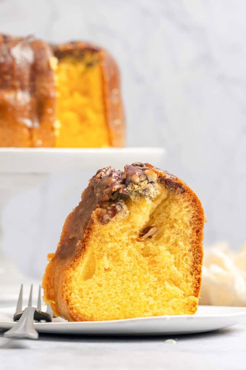 A slice of rum cake with a pecan topping and glaze sits on a plate with a fork. The full bundt cake is on a stand behind it.