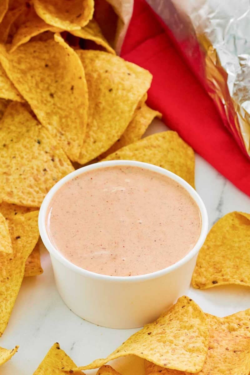 A close-up of a cup of creamy chipotle adobo ranch dip, with tortilla chips spilling out from a bag in the background. The image highlights the smooth texture of the dip, perfect for snacking.
