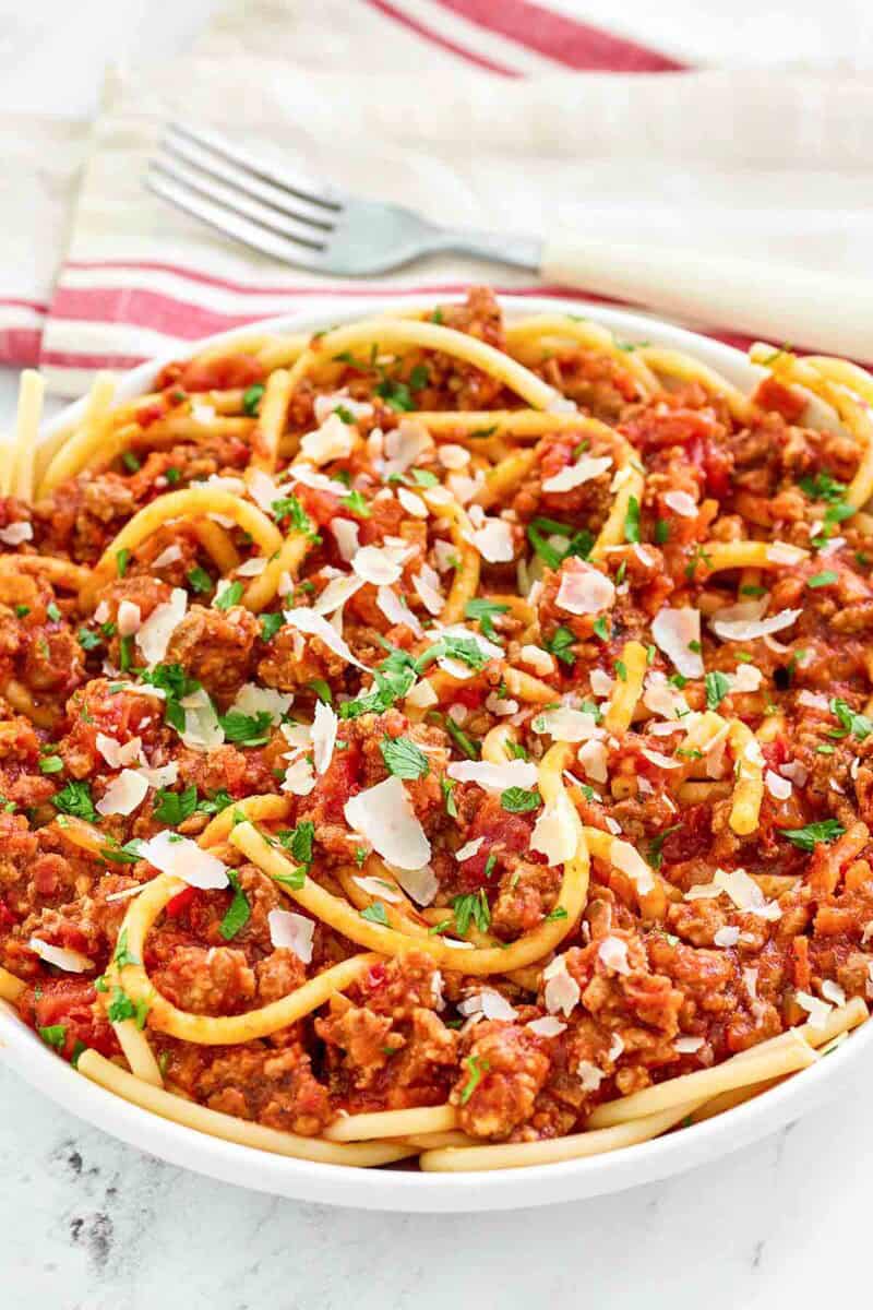 A close-up of a bowl of pasta with Olive Garden's spicy three-meat sauce. The rich, chunky texture of the meat sauce is prominent as it coats the pasta, which is topped with shaved parmesan and fresh parsley.