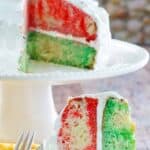 Homemade Jell-O poke cake with whipped cream frosting on a cake stand and a slice on a plate.