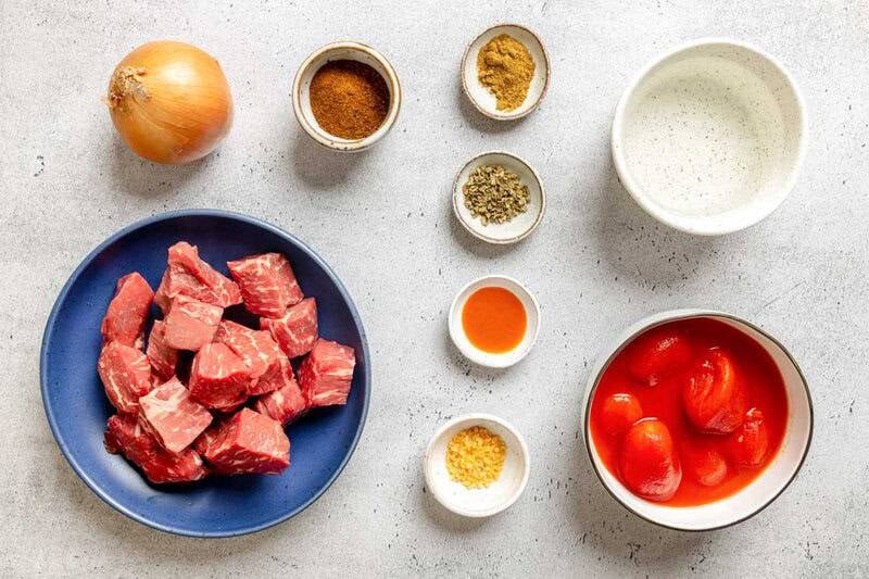 An overhead view of ingredients for chili: cubed beef, onion, tomatoes, water, chili powder, cumin, oregano, garlic, and hot sauce.