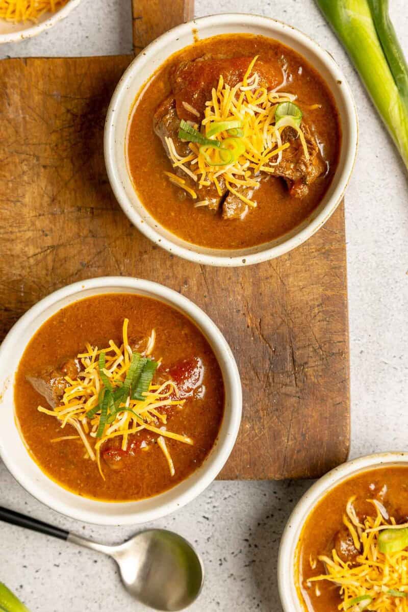 An overhead shot of two bowls of Pedernales River Chili, topped with shredded cheese and green onions, sitting on a wooden board.