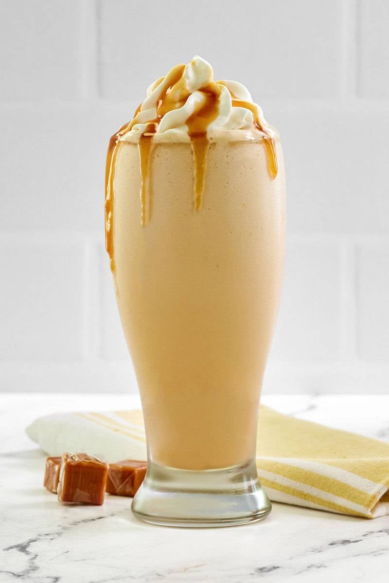 A tall glass of a blended McDonald's caramel frappe, topped with whipped cream and caramel drizzle, with caramel candies beside it.
