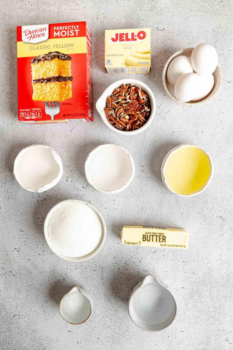 Ingredients for rum cake on a table: yellow cake mix, vanilla pudding, eggs, pecans, sugar, butter, and liquids in bowls.