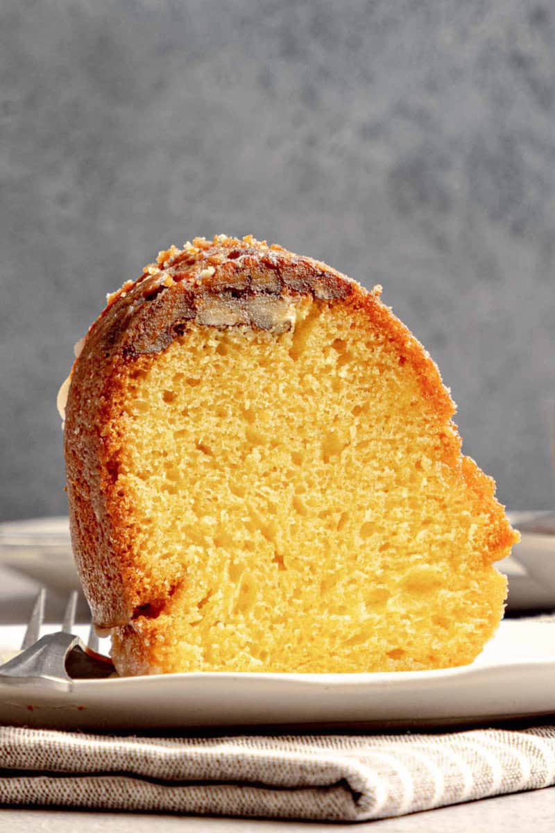 A close-up side view of a single slice of New Orleans rum cake, showing its moist, golden crumb and a pecan-studded glazed crust.