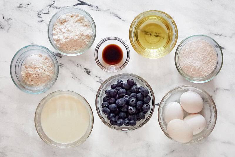 Overhead view of ingredients for blueberry muffins in glass bowls: flour, oil, milk, eggs, vanilla, and fresh blueberries.