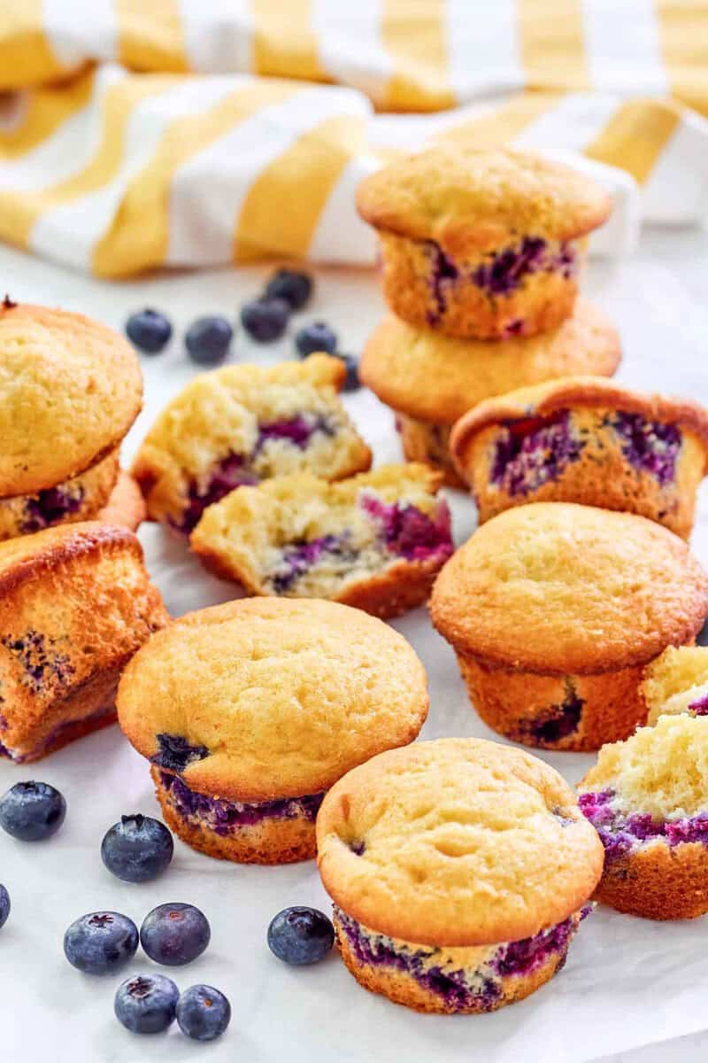 A cluster of golden-brown blueberry muffins, some stacked and one on its side, revealing the baked blueberries.