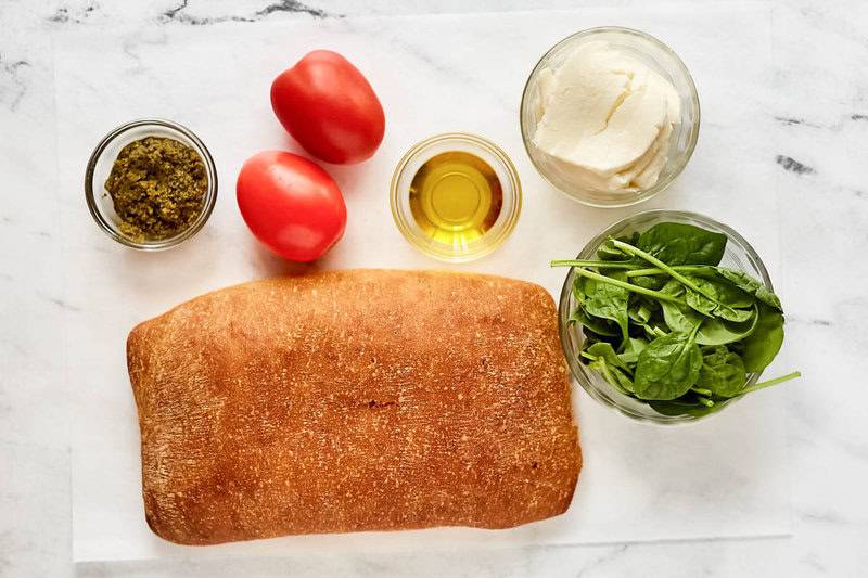 Ingredients for Starbucks Tomato Mozzarella sandwich: focaccia, tomatoes, mozzarella, spinach, pesto, and olive oil on a table.