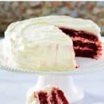 A homemade Waldorf Astoria red velvet cake on a a cake stand and a slice on a plate in front of it.