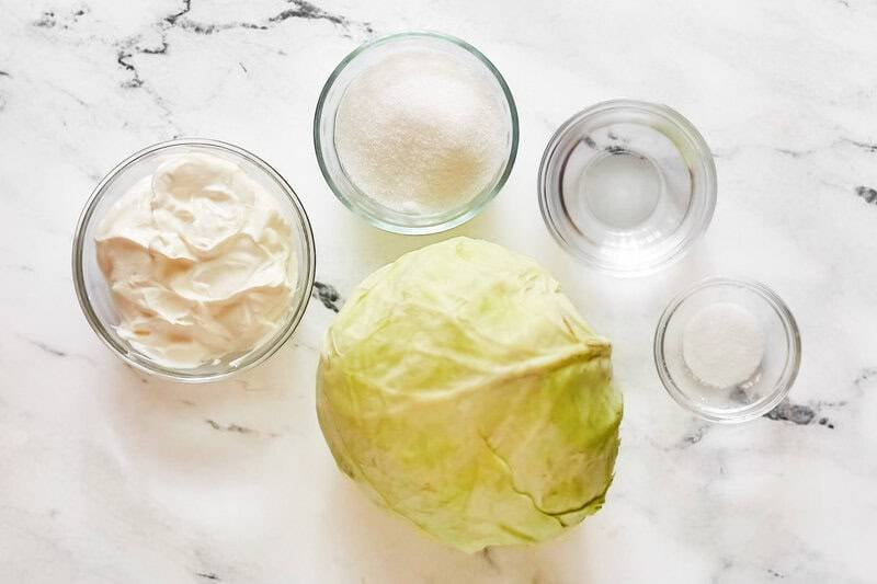 An overhead view of the simple ingredients: a head of cabbage, sour cream, sugar, vinegar, and salt in small glass bowls.