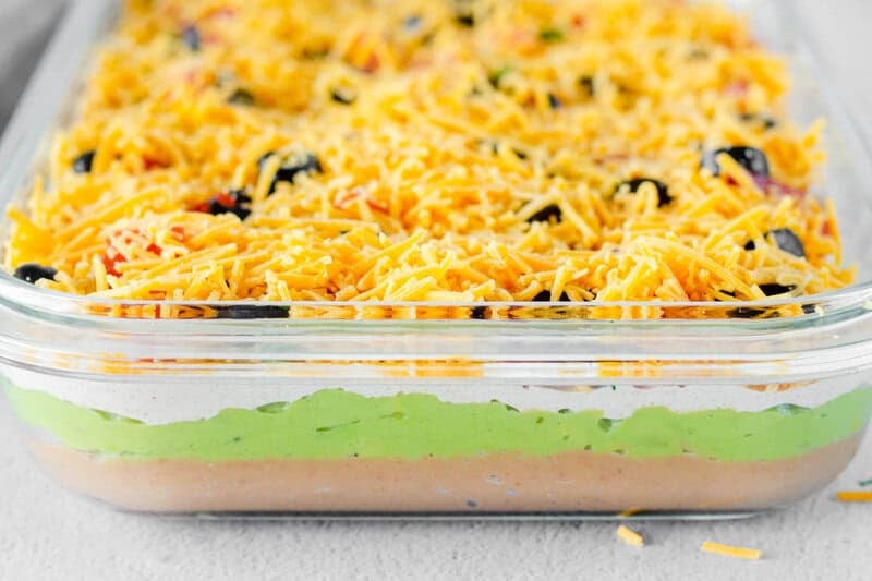 Side view of a glass dish filled with 7-layer dip, revealing distinct layers of beans, guacamole, sour cream, and cheese.