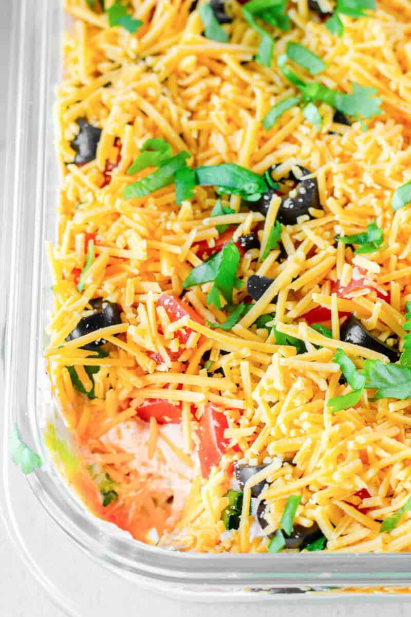 7-layer dip topped with shredded cheddar, chopped tomatoes, black olives, and green onions in a glass dish