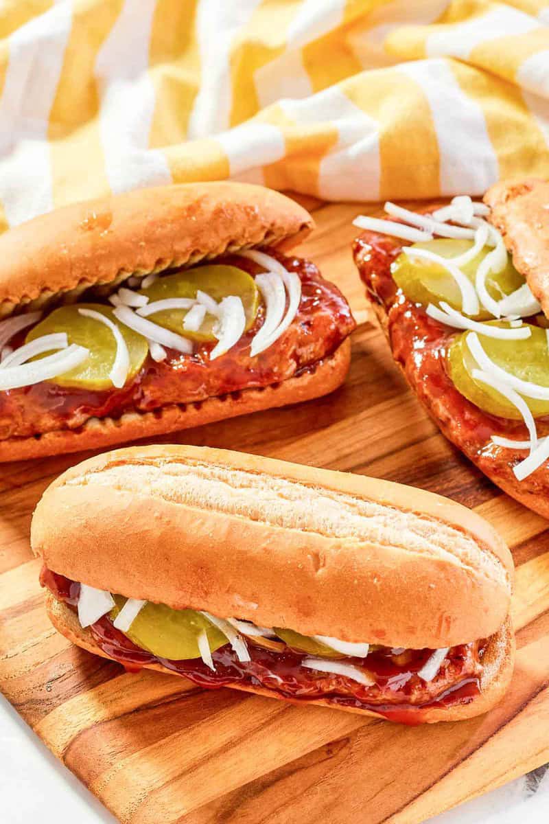 Three homemade McRib sandwiches on a wooden board with pickles and onions visible under the top bun.