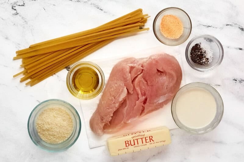 uncooked fettuccine, raw chicken breast, butter, cream, parmesan cheese, spices, and oil on a white table.