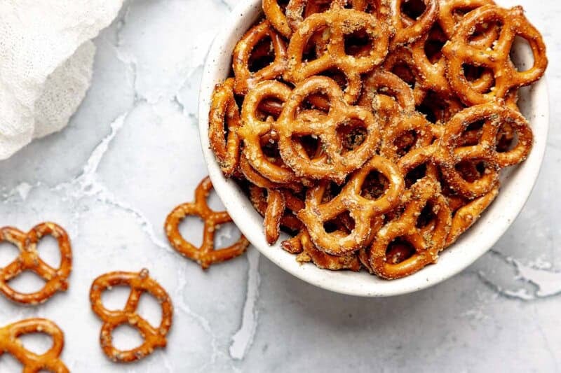 A bowl of homemade ranch pretzels and a few pretzels scattered by it.