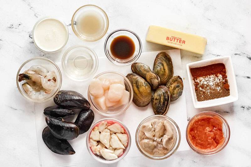 Overhead view of ingredients: fresh shrimp, scallops, mussels, clams, crab, spices, butter, and cream on a white surface.