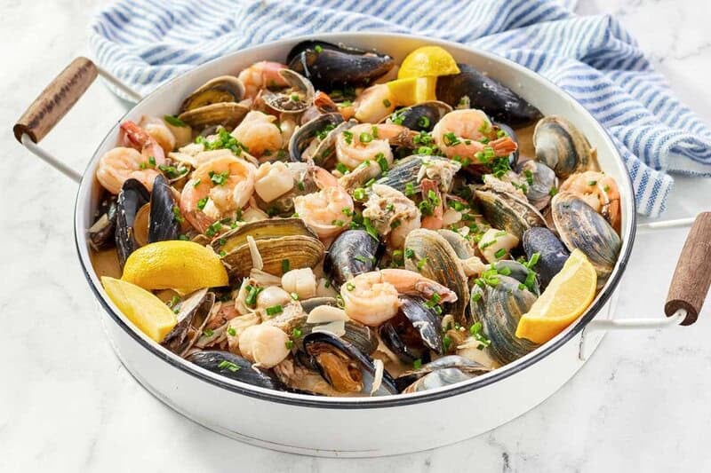 A close-up overhead view of a rich seafood pan roast with clams, shrimp, and mussels in a savory broth with lemon wedges.