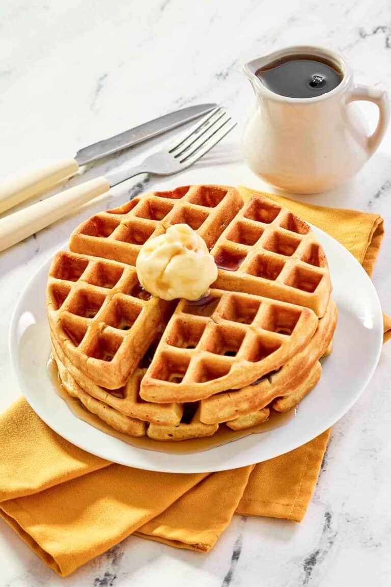 Three homemade waffles stacked on a plate and topped with butter and syrup.
