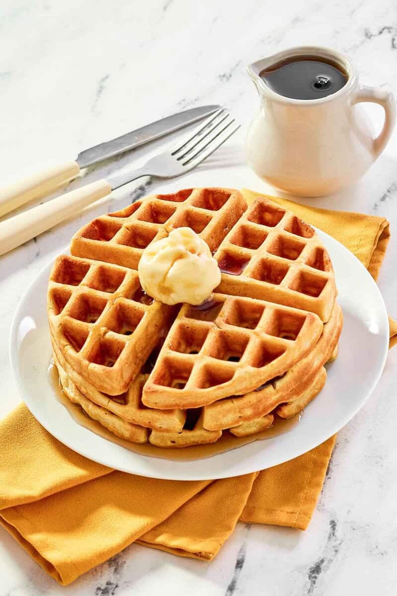 Three homemade waffles stacked on a plate and topped with butter and syrup.