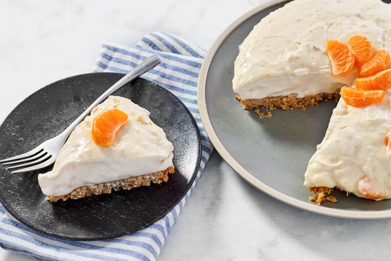 A slice of creamy cheesecake with mandarin oranges on a plate next to the cheesecake.