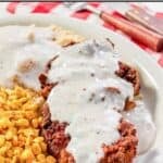 A crispy chicken fried steak topped with white gravy on a plate with mashed potatoes and corn.